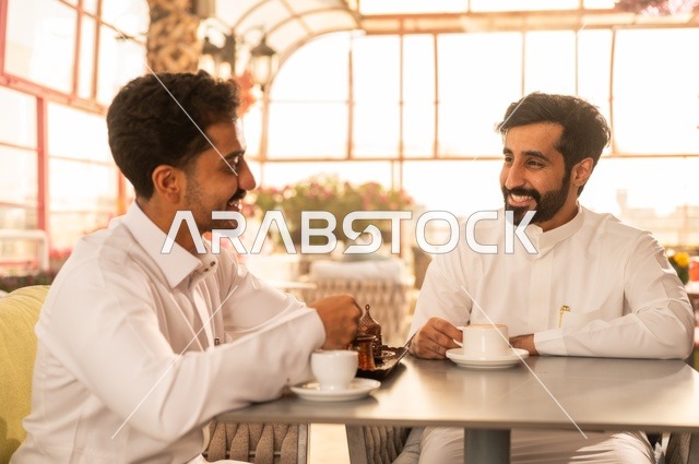 Sharing fun times with friends, building successful social relationships, exchanging conversations and news, enjoying hot coffee, two Saudi Arabian Gulf friends wearing traditional dress sitting in one of the luxurious cafes in the Kingdom