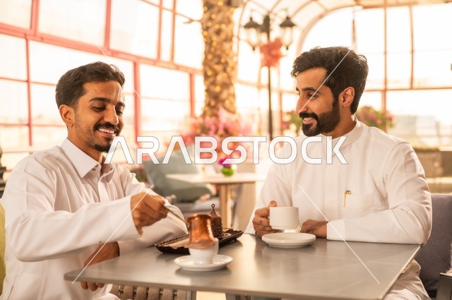 Building successful social relationships, exchanging conversations and news, sharing fun times with friends, enjoying hot coffee, two Saudi Arabian Gulf friends wearing traditional dress sitting in one of the luxurious cafes in the Kingdom