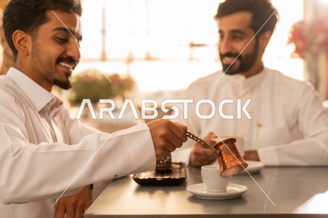 بناء علاقات اجتماعية ناجحة, تبادل الأحاديث والاخبار, مشاركة الأصدقاء الأوقات الممتعة, الاستمتاع بتناول القهوة الساخنة, صديقان عربيان خليجيان سعوديان يرتديان الزي التقليدي يجلسانفي إحدى الكافيهات الفاخرة في المملكة