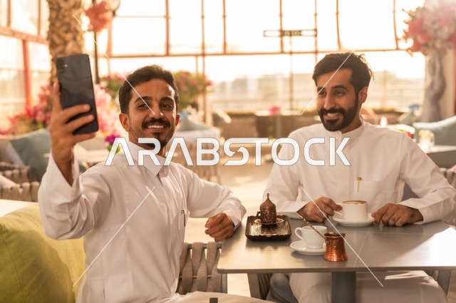 التقاط صورة سيلفي تذكارية باستخدام الهاتف المحمول، مشاركة الأصدقاء الأوقات الممتعة, بناء علاقات اجتماعية ناجحة, تبادل الأحاديث والاخبار, الاستمتاع بتناول القهوة الساخنة, صديقان عربيان خليجيان سعوديان يرتديان الزي التقليدي يجلسان في إحدى الكافيهات الفاخرة في المملكة