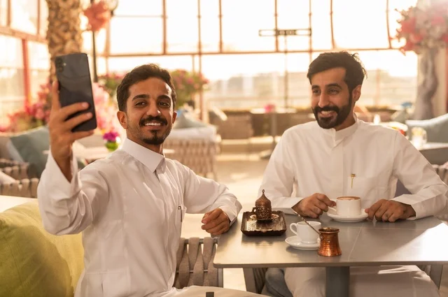 التقاط صورة سيلفي تذكارية باستخدام الهاتف المحمول، مشاركة الأصدقاء الأوقات الممتعة, بناء علاقات اجتماعية ناجحة, تبادل الأحاديث والاخبار, الاستمتاع بتناول القهوة الساخنة, صديقان عربيان خليجيان سعوديان يرتديان الزي التقليدي يجلسان في إحدى الكافيهات الفاخرة في المملكة