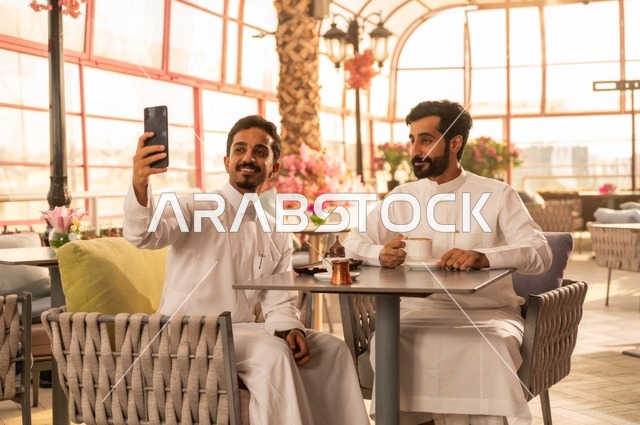 Taking a selfie with your mobile phone, sharing fun times with friends, building successful social relationships, exchanging conversations and news, enjoying hot coffee, two Saudi Arabian Gulf friends wearing traditional clothing sitting in one of the luxurious cafes in the Kingdom
