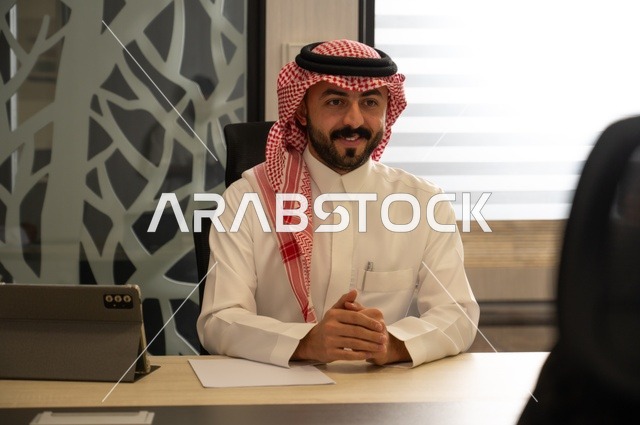 Administrative office jobs for youth in Saudi Arabia, integrating work with technological advancements, the concept of remote work via the internet, an Arab Gulf Saudi man wearing traditional attire and a headscarf sitting behind a desk with a tablet in front of him.