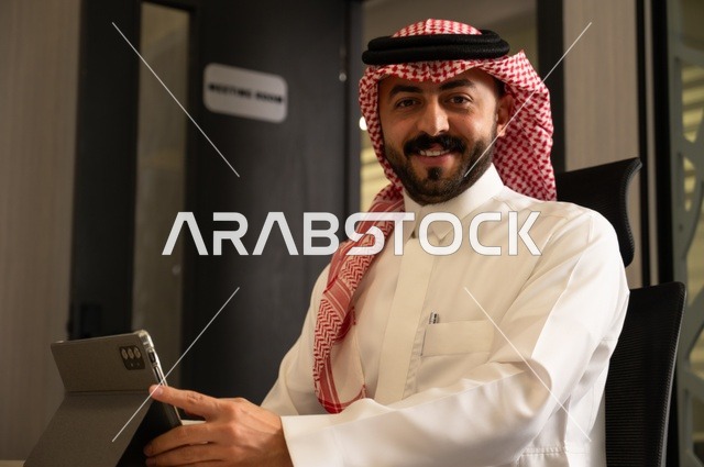 مهن ووظائف إدارية مكتبية للشباب في السعودية, دمج العمل مع التقنيات التكنولوجية، مفهوم العمل عن بعد عبر الانترنت، رجل عربي خليجي سعودي يرتدي الثوب التقليدي والشماغ يجلس خلف المكتب وأمامه جهاز التابلت