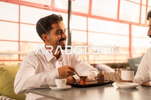 مشاركة الأصدقاء الأوقات الممتعة, بناء علاقات اجتماعية ناجحة, تبادل الأحاديث والاخبار, الاستمتاع بتناول القهوة الساخنة, صديقان عربيان خليجيان سعوديان يرتديان الزي التقليدي يجلسان في إحدى الكافيهات الفاخرة في المملكة