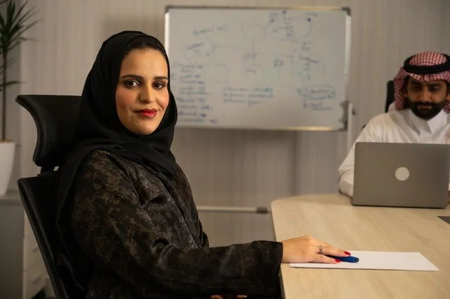 Data analysis results, a Saudi Arabian Gulf Arab woman wearing a hijab and an abaya sits behind the table taking notes on paper, a female office job in Gulf Arab companies.