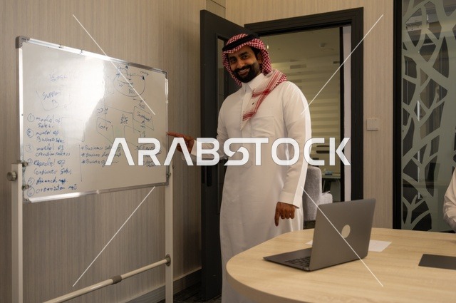 Using the whiteboard to clarify the processes and procedures followed in the company, teamwork and team spirit, discussing work plans, using the laptop to take notes, professionalism and dedication to achievement, a Saudi Gulf Arab team in a meeting inside the office, ensuring a clear and comprehensive understanding of the work.
