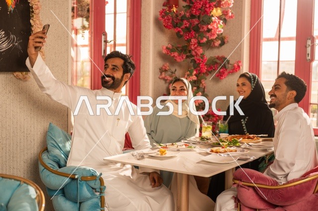 Taking a commemorative selfie using a mobile phone, sharing enjoyable moments with friends, enjoying lunch at a restaurant, a family of Saudi Gulf Arabs sitting at the table in one of the upscale restaurants and eating with gestures of happiness and joy.