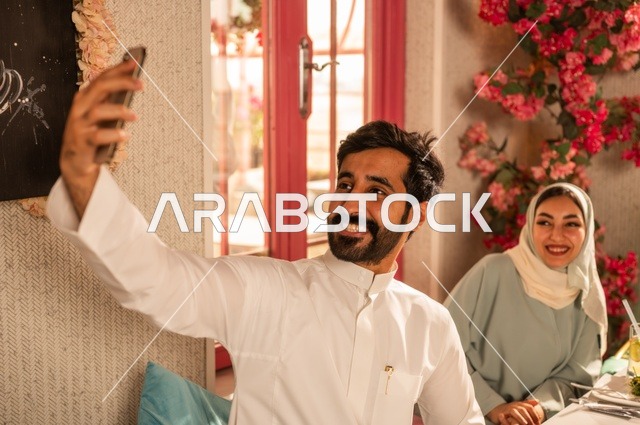 Taking a commemorative selfie using a mobile phone, sharing enjoyable moments with friends, enjoying lunch at a restaurant, a family of Saudi Gulf Arabs sitting at the table in one of the upscale restaurants and eating with gestures of happiness and joy.