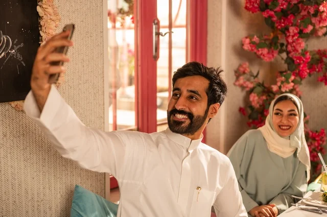 التقاط صورة سيلفي تذكارية باستخدام الهاتف المحمول, مشاركة الأصدقاء الأوقات الممتعة, الاستمتاع بتناول الغداء في المطعم, عائلة عرب خليجيون سعوديون يجلسون على المائدة في إحدى المطاعم الراقية ويقومون بتناول الطعام بإيماءات السعادة والسرور