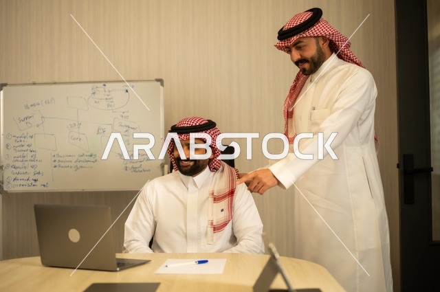 تبادل الخبرات والافكار، دراسة وتحليل المشاريع الجديدة، استخدام جهاز تقني حديث، الوحدة والعمل الجماعي، تطوير العمل وحل المشكلات، رجل عربي خليجي سعودي يرتدي الشماغ والثوب التقليدي يقوم بالعمل برفقة مديره، مهنة ووظيفة سعودية مكتبية