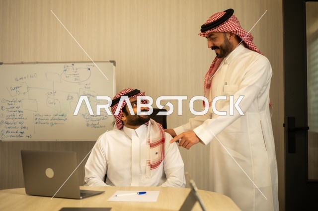 تبادل الخبرات والافكار، دراسة وتحليل المشاريع الجديدة، استخدام جهاز تقني حديث، الوحدة والعمل الجماعي، تطوير العمل وحل المشكلات، رجل عربي خليجي سعودي يرتدي الشماغ والثوب التقليدي يقوم بالعمل برفقة مديره، مهنة ووظيفة سعودية مكتبية