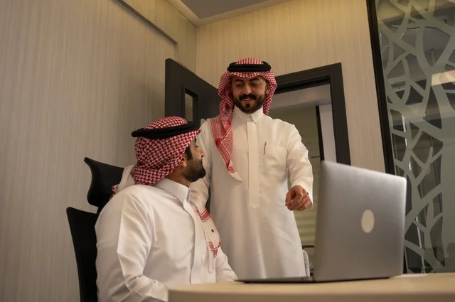 Exchange of experiences and ideas, studying and analyzing new projects, using modern technical devices, unity and teamwork, developing work and solving problems, an Arab Gulf Saudi man wearing the traditional headscarf and thobe working alongside his manager, a Saudi office profession and job.