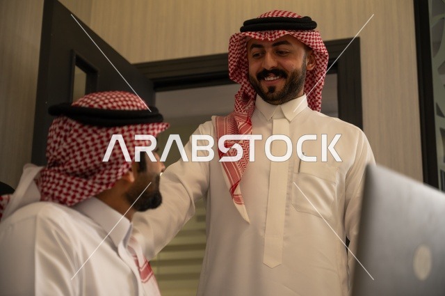 تبادل الخبرات والافكار، دراسة وتحليل المشاريع الجديدة، استخدام جهاز تقني حديث، الوحدة والعمل الجماعي، تطوير العمل وحل المشكلات، رجل عربي خليجي سعودي يرتدي الشماغ والثوب التقليدي يقوم بالعمل برفقة مديره، مهنة ووظيفة سعودية مكتبية