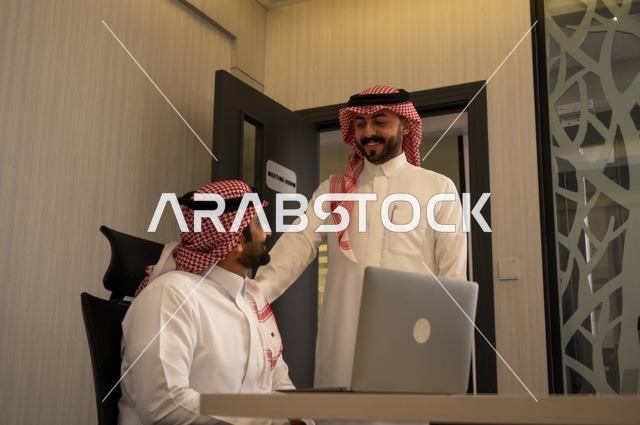 تبادل الخبرات والافكار، دراسة وتحليل المشاريع الجديدة، استخدام جهاز تقني حديث، الوحدة والعمل الجماعي، تطوير العمل وحل المشكلات، رجل عربي خليجي سعودي يرتدي الشماغ والثوب التقليدي يقوم بالعمل برفقة مديره، مهنة ووظيفة سعودية مكتبية
