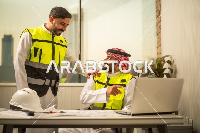 الشركات المختصة بورشات البناء والمقاولات، تصميم وتخطيط المخططات المعمارية عبر الحاسوب المحمول، متابعة تنفيذ الخطط الهندسية، رجلان مهندسان عربيان خليجيان سعوديان يرتدي زي العمل يقومان بالمناقشة وتبادل الخبرات، استخدام جهاز تقني حديث