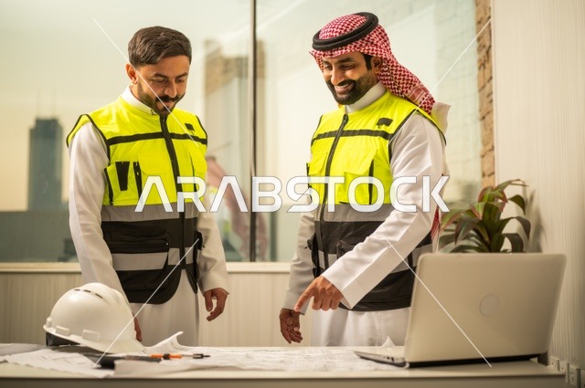 الشركات المختصة بورشات البناء والمقاولات، تصميم وتخطيط المخططات المعمارية عبر الحاسوب المحمول، متابعة تنفيذ الخطط الهندسية، رجلان مهندسان عربيان خليجيان سعوديان يرتدي زي العمل يقومان بالمناقشة وتبادل الخبرات، استخدام جهاز تقني حديث