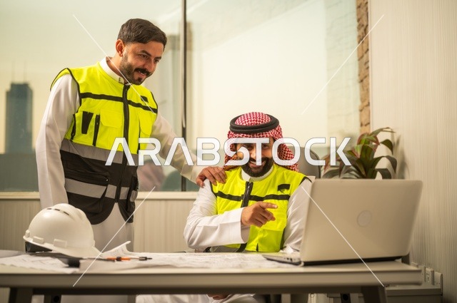 الشركات المختصة بورشات البناء والمقاولات، تصميم وتخطيط المخططات المعمارية عبر الحاسوب المحمول، متابعة تنفيذ الخطط الهندسية، رجلان مهندسان عربيان خليجيان سعوديان يرتدي زي العمل يقومان بالمناقشة وتبادل الخبرات، استخدام جهاز تقني حديث