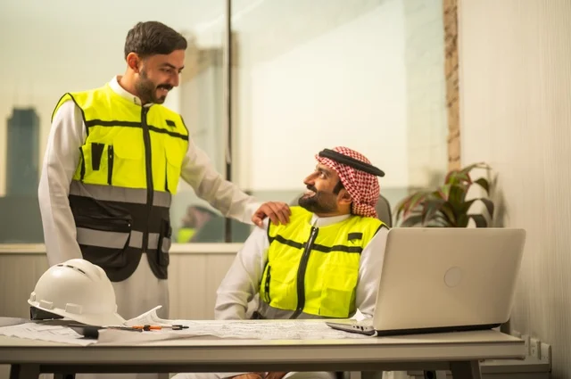 الشركات المختصة بورشات البناء والمقاولات، تصميم وتخطيط المخططات المعمارية عبر الحاسوب المحمول، متابعة تنفيذ الخطط الهندسية، رجلان مهندسان عربيان خليجيان سعوديان يرتدي زي العمل يقومان بالمناقشة وتبادل الخبرات، استخدام جهاز تقني حديث