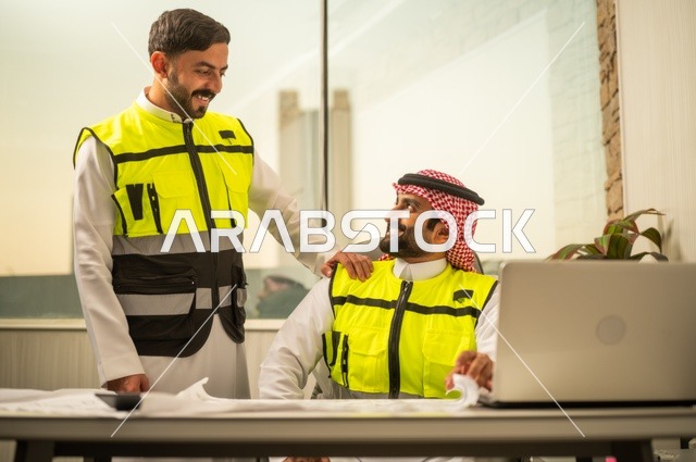 الشركات المختصة بورشات البناء والمقاولات، تصميم وتخطيط المخططات المعمارية عبر الحاسوب المحمول، متابعة تنفيذ الخطط الهندسية، رجلان مهندسان عربيان خليجيان سعوديان يرتدي زي العمل يقومان بالمناقشة وتبادل الخبرات، استخدام جهاز تقني حديث