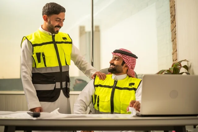 الشركات المختصة بورشات البناء والمقاولات، تصميم وتخطيط المخططات المعمارية عبر الحاسوب المحمول، متابعة تنفيذ الخطط الهندسية، رجلان مهندسان عربيان خليجيان سعوديان يرتدي زي العمل يقومان بالمناقشة وتبادل الخبرات، استخدام جهاز تقني حديث