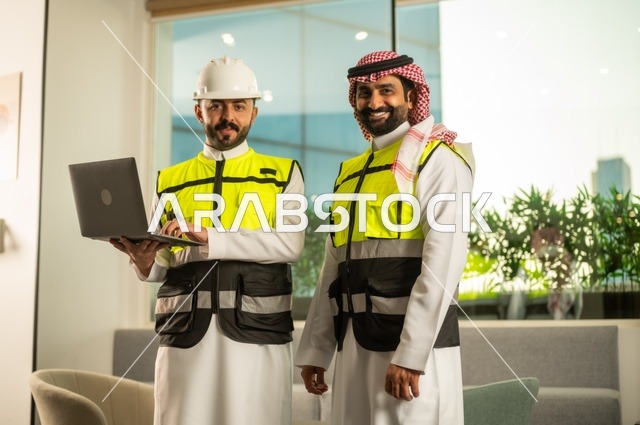 الشركات المختصة بورشات البناء والمقاولات، تصميم وتخطيط المخططات المعمارية عبر الحاسوب المحمول، متابعة تنفيذ الخطط الهندسية، رجلان مهندسان عربيان خليجيان سعوديان يرتدي زي العمل يقومان بالمناقشة وتبادل الخبرات، استخدام جهاز تقني حديث
