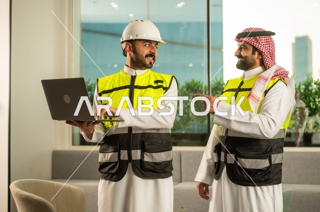 الشركات المختصة بورشات البناء والمقاولات، تصميم وتخطيط المخططات المعمارية عبر الحاسوب المحمول، متابعة تنفيذ الخطط الهندسية، رجلان مهندسان عربيان خليجيان سعوديان يرتدي زي العمل يقومان بالمناقشة وتبادل الخبرات، استخدام جهاز تقني حديث