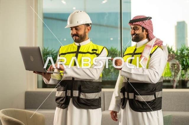 الشركات المختصة بورشات البناء والمقاولات، تصميم وتخطيط المخططات المعمارية عبر الحاسوب المحمول، متابعة تنفيذ الخطط الهندسية، رجلان مهندسان عربيان خليجيان سعوديان يرتدي زي العمل يقومان بالمناقشة وتبادل الخبرات، استخدام جهاز تقني حديث