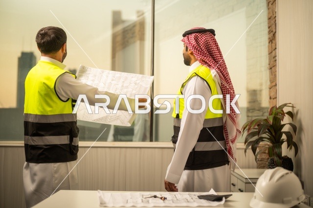 متابعة تنفيذ الخطط الهندسية، الشركات المختصة بورشات البناء والمقاولات، التخطيط ووضع اساسيات المخططات المعمارية، مهندسان عربيان خليجيان سعوديان يرتديان سترة الحماية يعملان على مناقشة مخطط الإنشاء وتحديد القياسات بدقة