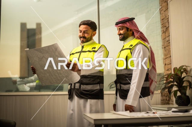 متابعة تنفيذ الخطط الهندسية، الشركات المختصة بورشات البناء والمقاولات، التخطيط ووضع اساسيات المخططات المعمارية، مهندسان عربيان خليجيان سعوديان يرتديان سترة الحماية يعملان على مناقشة مخطط الإنشاء وتحديد القياسات بدقة