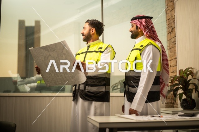 متابعة تنفيذ الخطط الهندسية، الشركات المختصة بورشات البناء والمقاولات، التخطيط ووضع اساسيات المخططات المعمارية، مهندسان عربيان خليجيان سعوديان يرتديان سترة الحماية يعملان على مناقشة مخطط الإنشاء وتحديد القياسات بدقة