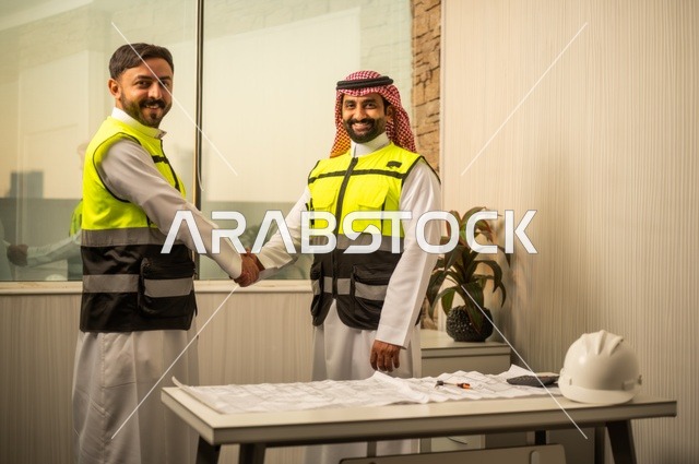 متابعة تنفيذ الخطط الهندسية، الشركات المختصة بورشات البناء والمقاولات، التخطيط ووضع اساسيات المخططات المعمارية، مهندسان عربيان خليجيان سعوديان يرتديان سترة الحماية يعملان على مناقشة مخطط الإنشاء وتحديد القياسات بدقة