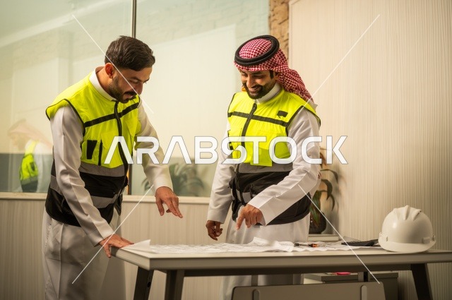 متابعة تنفيذ الخطط الهندسية، الشركات المختصة بورشات البناء والمقاولات، التخطيط ووضع اساسيات المخططات المعمارية، مهندسان عربيان خليجيان سعوديان يرتديان سترة الحماية يعملان على مناقشة مخطط الإنشاء وتحديد القياسات بدقة