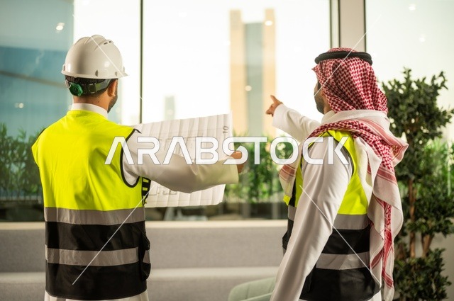 متابعة تنفيذ الخطط الهندسية، الشركات المختصة بورشات البناء والمقاولات، التخطيط ووضع اساسيات المخططات المعمارية، مهندسان عربيان خليجيان سعوديان يرتديان سترة الحماية يعملان على مناقشة مخطط الإنشاء وتحديد القياسات بدقة