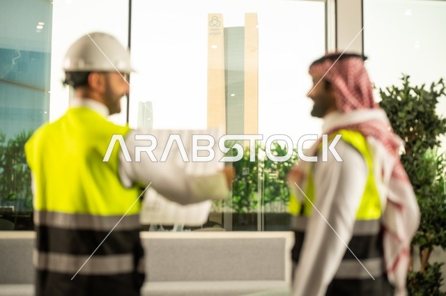 Monitoring the implementation of engineering plans, companies specialized in construction workshops and contracting, planning and establishing the fundamentals of architectural designs, two Saudi Gulf Arab engineers wearing safety vests are discussing the construction plan and accurately determining the measurements.