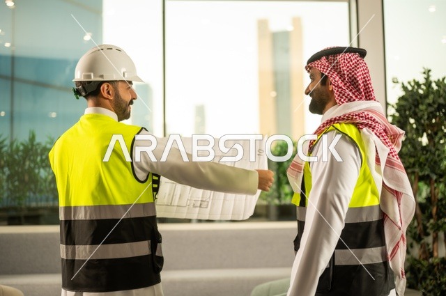 Monitoring the implementation of engineering plans, companies specialized in construction workshops and contracting, planning and establishing the fundamentals of architectural designs, two Saudi Gulf Arab engineers wearing safety vests are discussing the construction plan and accurately determining the measurements.