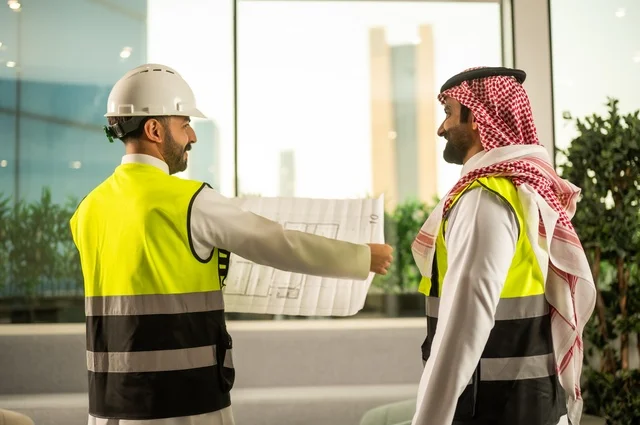 Monitoring the implementation of engineering plans, companies specialized in construction workshops and contracting, planning and establishing the fundamentals of architectural designs, two Saudi Gulf Arab engineers wearing safety vests are discussing the construction plan and accurately determining the measurements.