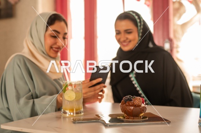 قضاء أوقات ممتعة مع الأصدقاء، النقاش والحوار في الأجواء الهادئة، مشاركة المشروبات الباردة والحلوى، صديقتان عربيتان خليجيتان سعوديتان تجلسان على الأريكة في إحدى المقاهي الرقية تقومان بمشاهده الهاتف المحمول، استخدام جهاز تقني حديث