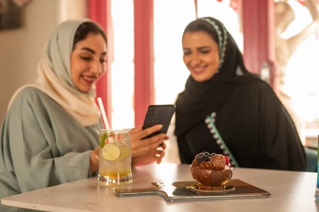 قضاء أوقات ممتعة مع الأصدقاء، النقاش والحوار في الأجواء الهادئة، مشاركة المشروبات الباردة والحلوى، صديقتان عربيتان خليجيتان سعوديتان تجلسان على الأريكة في إحدى المقاهي الرقية تقومان بمشاهده الهاتف المحمول، استخدام جهاز تقني حديث