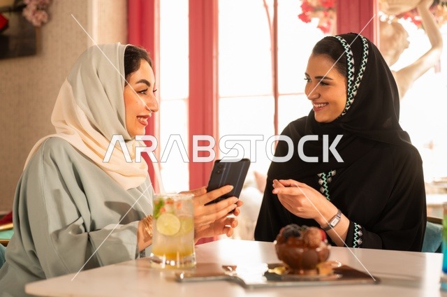 قضاء أوقات ممتعة مع الأصدقاء، النقاش والحوار في الأجواء الهادئة، مشاركة المشروبات الباردة والحلوى، صديقتان عربيتان خليجيتان سعوديتان تجلسان على الأريكة في إحدى المقاهي الرقية تقومان بمشاهده الهاتف المحمول، استخدام جهاز تقني حديث