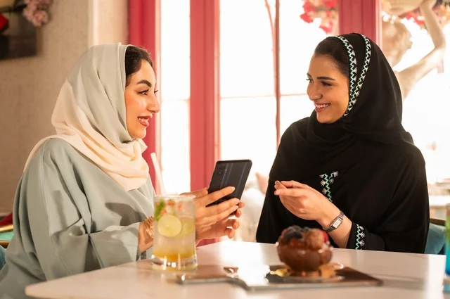 Spending enjoyable times with friends, discussing and conversing in a calm atmosphere, sharing cold drinks and sweets, two Arab Gulf friends from Saudi Arabia are sitting on a sofa in a luxurious café, watching on a mobile phone, using a modern technological device.