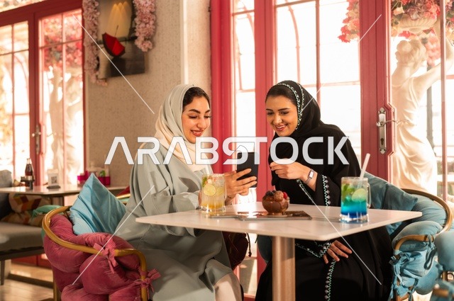 قضاء أوقات ممتعة مع الأصدقاء، النقاش والحوار في الأجواء الهادئة، مشاركة المشروبات الباردة والحلوى، صديقتان عربيتان خليجيتان سعوديتان تجلسان على الأريكة في إحدى المقاهي الرقية تقومان بمشاهده الهاتف المحمول، استخدام جهاز تقني حديث