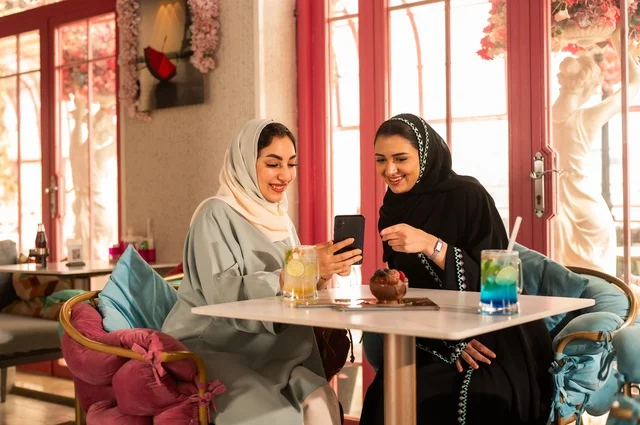 Spending enjoyable times with friends, discussing and conversing in a calm atmosphere, sharing cold drinks and sweets, two Arab Gulf friends from Saudi Arabia are sitting on a sofa in a luxurious café, watching on a mobile phone, using a modern technological device.