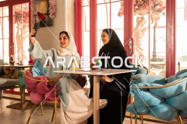 التقاط صورة سيلفي تذكارية باستخدام الهاتف المحمول، قضاء أوقات ممتعة مع الأصدقاء، النقاش والحوار في الأجواء الهادئة، مشاركة المشروبات الباردة والحلوى، صديقتان عربيتان خليجيتان سعوديتان تجلسان على الأريكة في إحدى المقاهي الراقية، استخدام جهاز تقني حديث