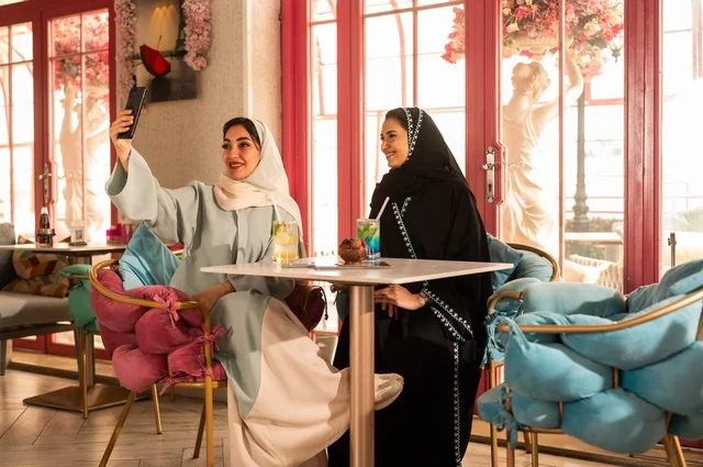 Taking a commemorative selfie using a mobile phone, spending enjoyable times with friends, discussing and conversing in a calm atmosphere, sharing cold drinks and sweets, two Arab Gulf Saudi friends sitting on a sofa in a high-end café, using modern technology.