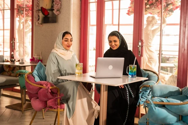 انجاز مهام العمل في الخارج، قضاء أوقات ممتعة مع الأصدقاء، النقاش والحوار في الأجواء الهادئة، مشاركة المشروبات الباردة والحلوى، صديقتان عربيتان خليجيتان سعوديتان تجلسان على الأريكة في إحدى المقاهي الرقية تقومان بمشاهده اللابتوب المحمول، استخدام جهاز تقني حديث