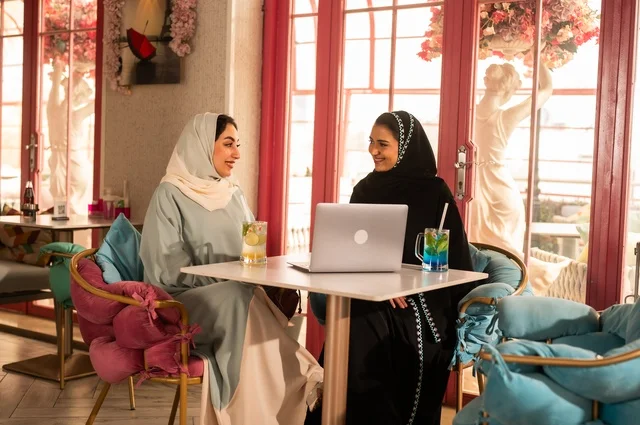 Completing work tasks abroad, spending enjoyable times with friends, discussing and conversing in calm atmospheres, sharing cold drinks and sweets, two Arab Gulf Saudi friends are sitting on a sofa in a luxurious café watching a laptop, using modern technology.