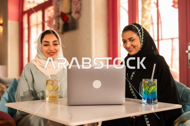 انجاز مهام العمل في الخارج، قضاء أوقات ممتعة مع الأصدقاء، النقاش والحوار في الأجواء الهادئة، مشاركة المشروبات الباردة والحلوى، صديقتان عربيتان خليجيتان سعوديتان تجلسان على الأريكة في إحدى المقاهي الرقية تقومان بمشاهده اللابتوب المحمول، استخدام جهاز تقني حديث