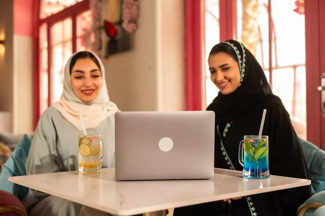 Completing work tasks abroad, spending enjoyable times with friends, discussing and conversing in calm atmospheres, sharing cold drinks and sweets, two Arab Gulf Saudi friends are sitting on a sofa in a luxurious café watching a laptop, using modern technology.