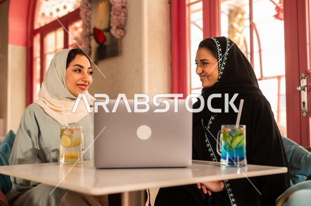 انجاز مهام العمل في الخارج، قضاء أوقات ممتعة مع الأصدقاء، النقاش والحوار في الأجواء الهادئة، مشاركة المشروبات الباردة والحلوى، صديقتان عربيتان خليجيتان سعوديتان تجلسان على الأريكة في إحدى المقاهي الرقية تقومان بمشاهده اللابتوب المحمول، استخدام جهاز تقني حديث